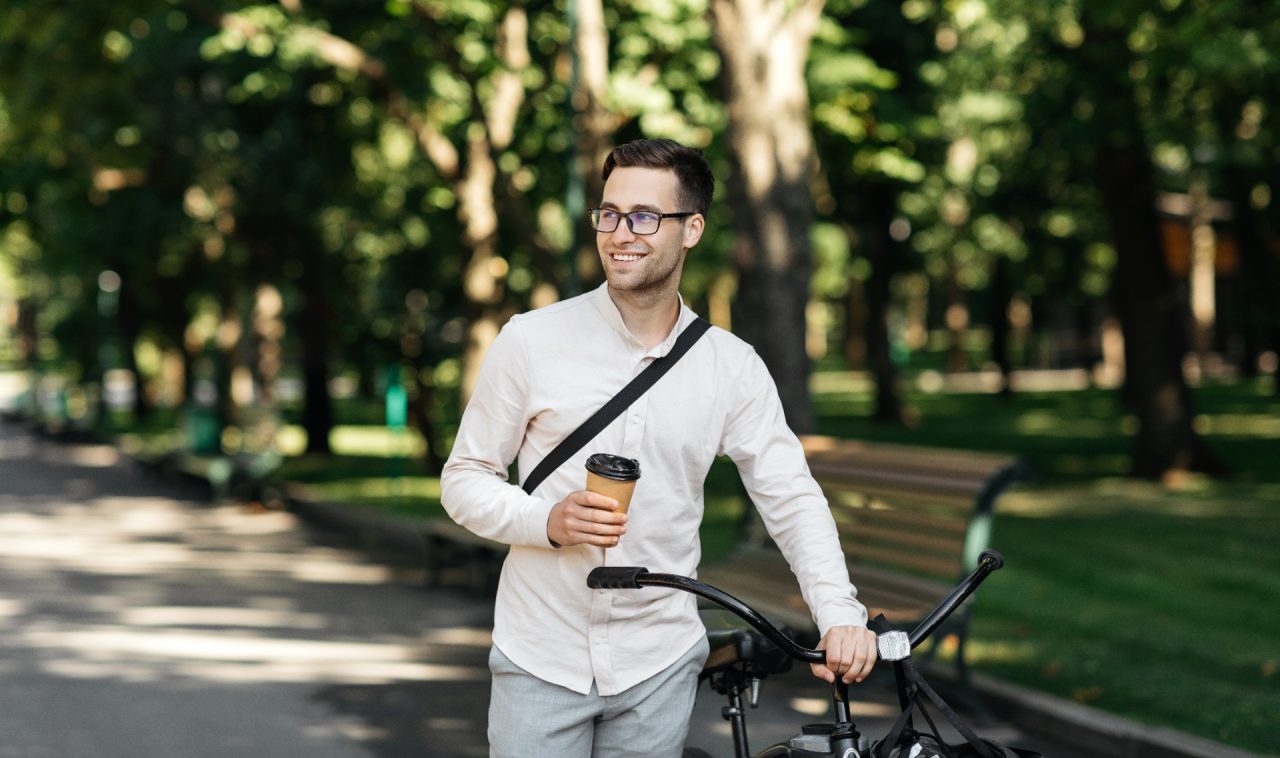 Covid-19 virus, urban eco transport and coffee takeaway on way to office. Happy young attractive male in formal clothes and glasses walks with bike and holds cup of hot drink in park, free space
