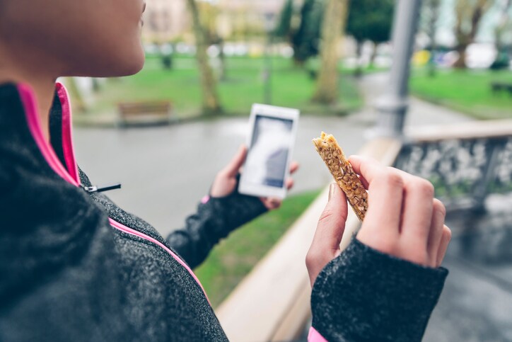 Woman eating cereal bar after training and holding smart phone