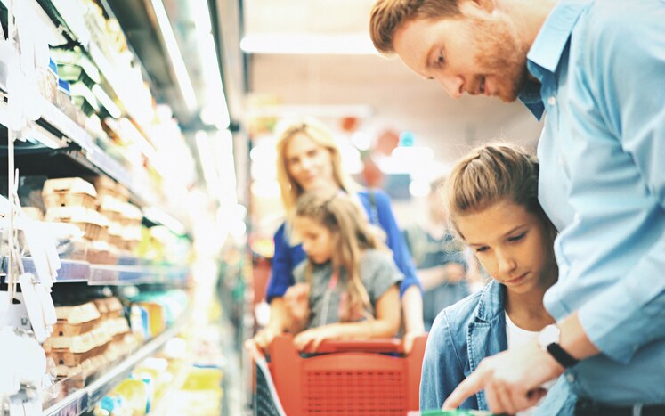 Closeup side view of young family with two daughters buying some frozen food in a supermarket. They are at refrigerated section and picking some frozen vegetables. Closeup on one of the daughters and father reading nutrition label on an unrecognizable product.