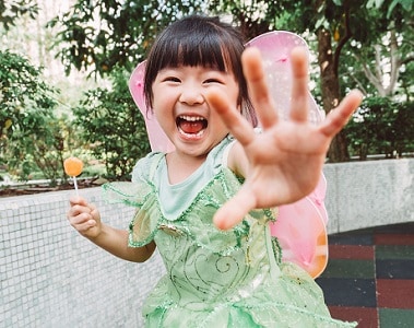 Lovely little girl in a fairy costume reaching out her hand to the camera and playing joyfully while holding a lollipop in her another hand in the playground