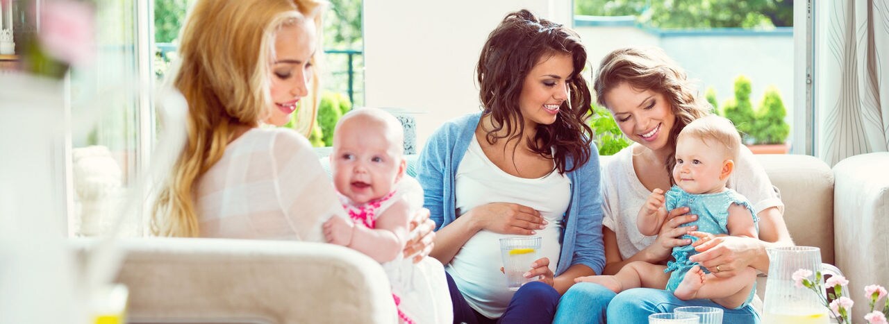 Two young women holding their baby girls on knee and talking wiht her pregnant friend.