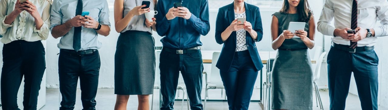 Cropped shot of multi-ethnic group of young business people using mobile devices together in modern office. Corporate business team with tablet and smart phones standing together in a line and having fun in their free time in the office.