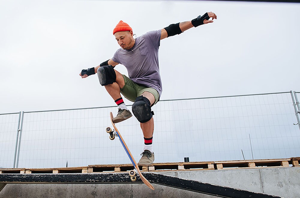 Agile mature skater in a watch cap doing tricks with a skateboard on a ramp in a skate park. Upward angle, hovering in the air with the board at an angle.