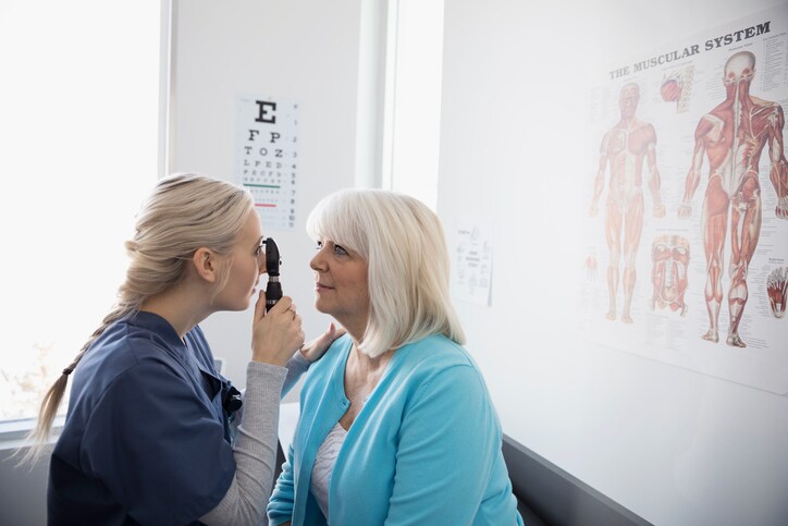 Female nurse examining eyes of senior patient with otoscope in clinic examination room