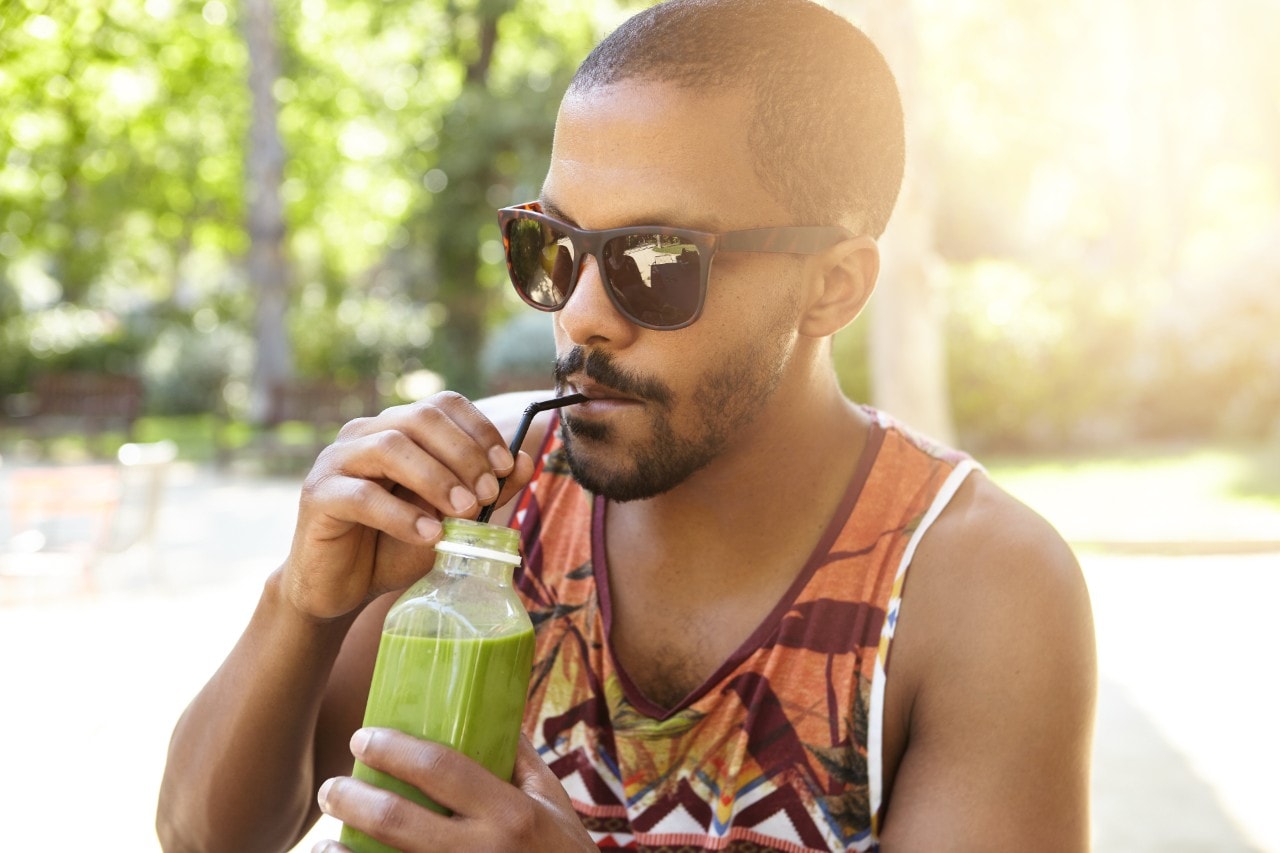 Healthy organic food. Half profile portrait of dark skinned guy drinking tasty fresh green smoothie using straw, sitting in shade on warm summer evening against blurred background of beautiful nature