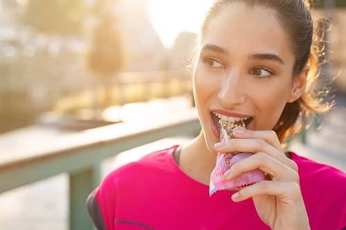 Athletic woman eating a protein bar. Closeup face of young sporty woman resting while biting a nutritive bar. Fitness beautiful woman eating a energy snack outdoor.