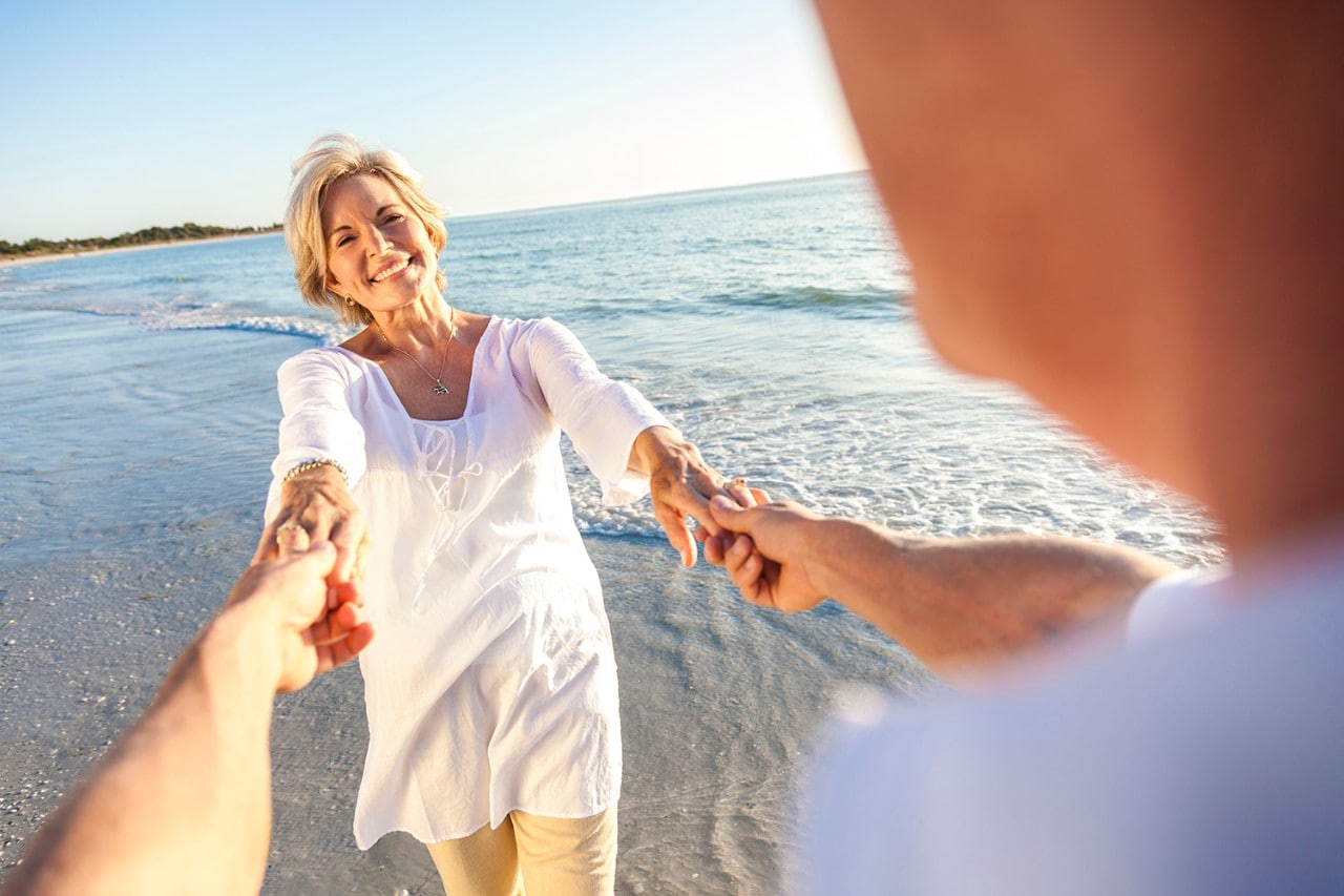 Happy senior man and woman couple walking or dancing and holding hands on a deserted tropical beach with bright clear blue sky