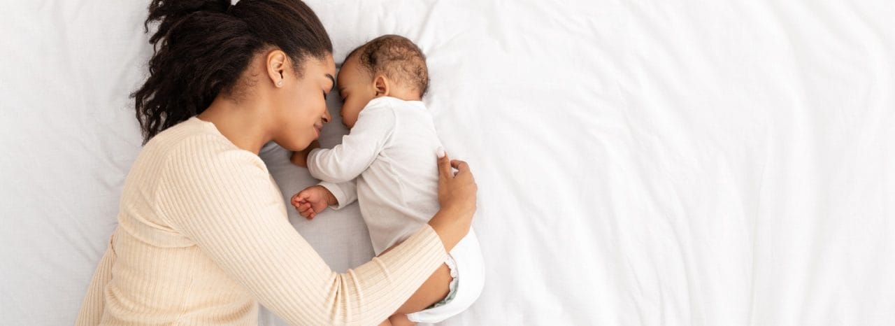 African Mother Hugging Sleeping Baby Lying In Bed Indoor, High Angle Shot. Loving Mom And Toddler Infant Napping Resting During Daytime Sleep. Panorama, Free Space For Text