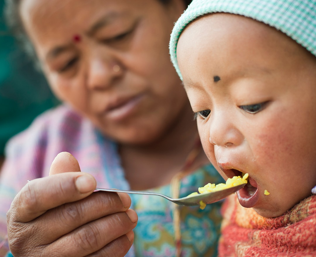 Outdoor image at day time of Asian woman feeding food to her grandson. The little child is opening his mouth wide to eat his lunch fed by his grandmother. Two people, Head and shoulders, horizontal composition and selective focus.