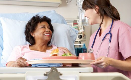 Smiling woman in hospital bed eating a meal provided by a nurse. 