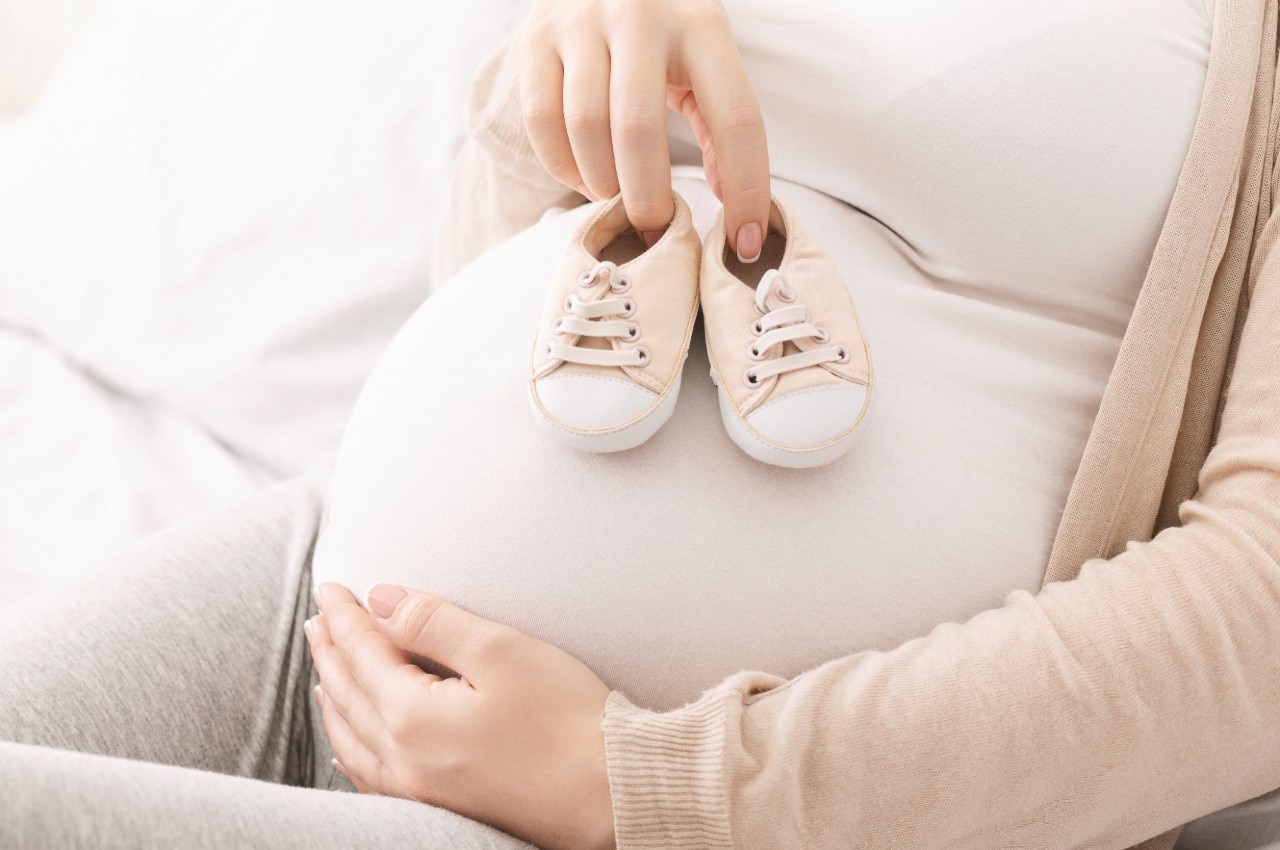Millennial woman playing with her belly, trying tiny baby shoes on it, close up, panorama with free space