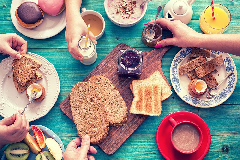 Foto de una mesa con bebidas, panes, frutas, cereales y mermeladas. Hay cuatro manos recogiendo algunos de estos artículos.