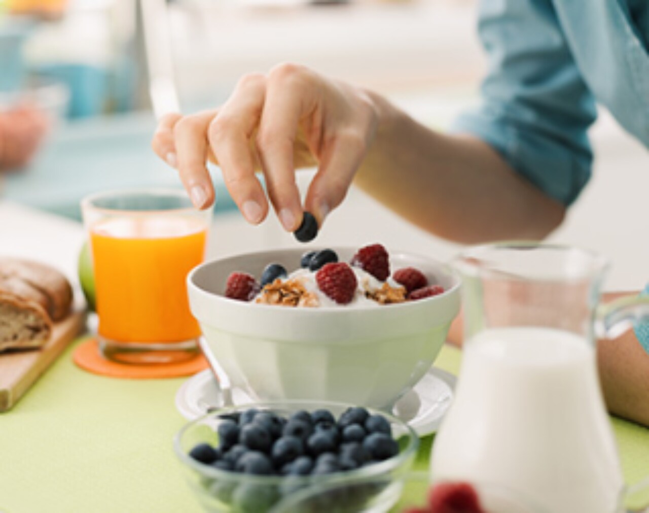 Foto de una mesa en la que hay tazones de fruta, un vaso con zumo de naranja y un bote con yogur. Hay una mano recogiendo frutas de uno de los tazones.