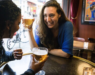 Foto de dos mujeres sonriendo, sentadas a una mesa en un bar. Están sosteniendo una copa con chop.