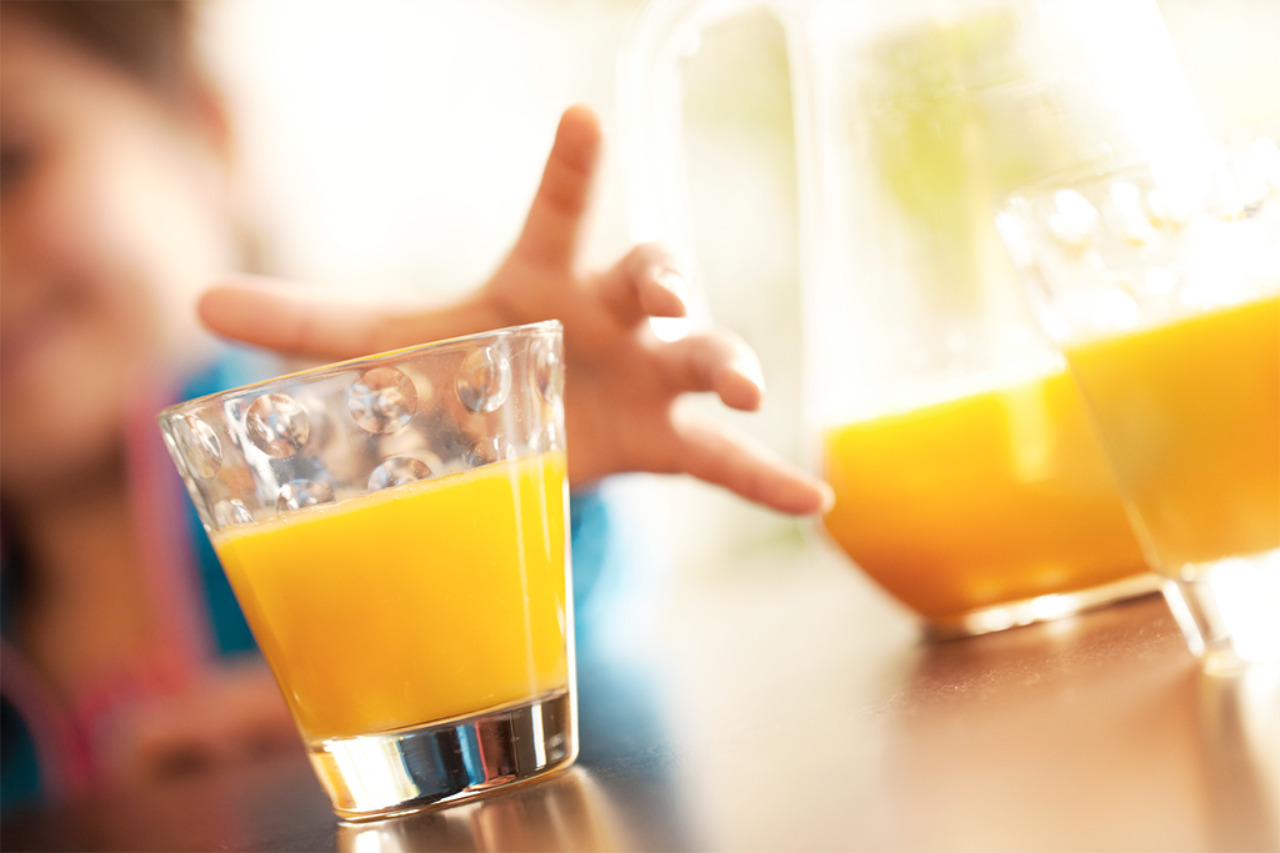 Foto de dos vasos y una jarra de jugo de naranja. En el fondo, se ve la mano de un niño preparándose para tomar uno de los vasos.