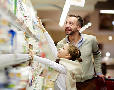 Foto de un hombre y una niña de pie frente a una estantería con productos desenfocados. La niña intenta alcanzar uno de los productos.