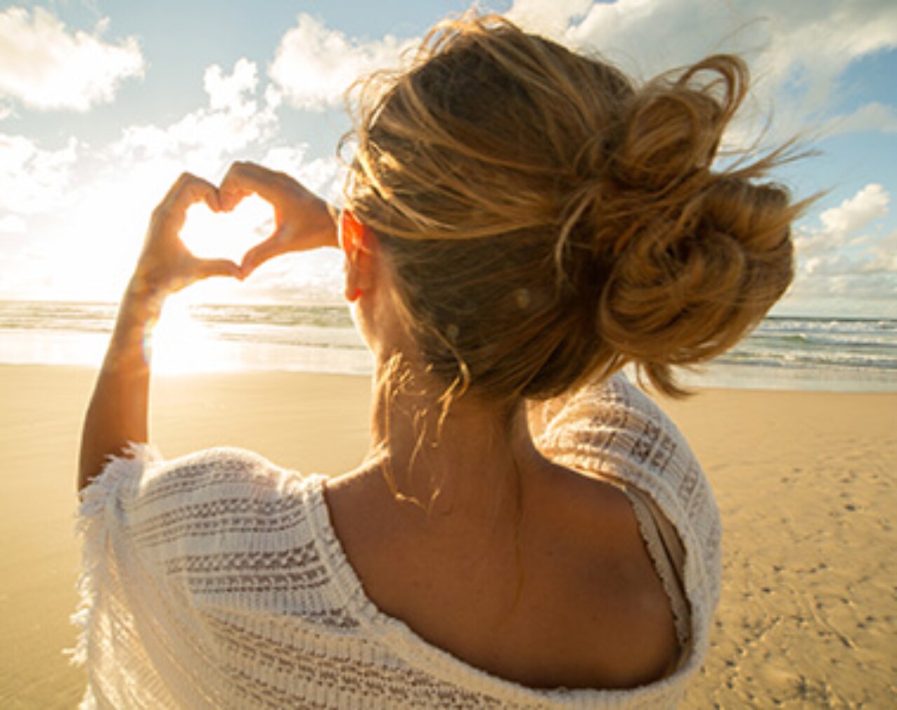 Foto de uma mulher de costas na praia. Ela está fazendo um coração com as duas mãos.