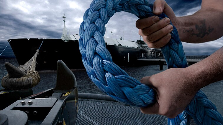 Foto de dos manos sosteniendo una cuerda gruesa. Al fondo, hay un barco en el mar.