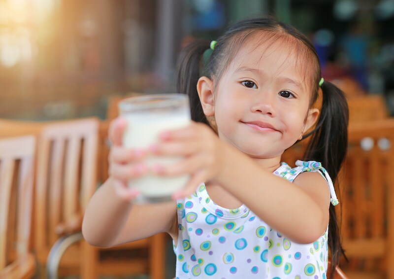 Foto de uma menina segurando, com as duas mãos, um copo com leite.