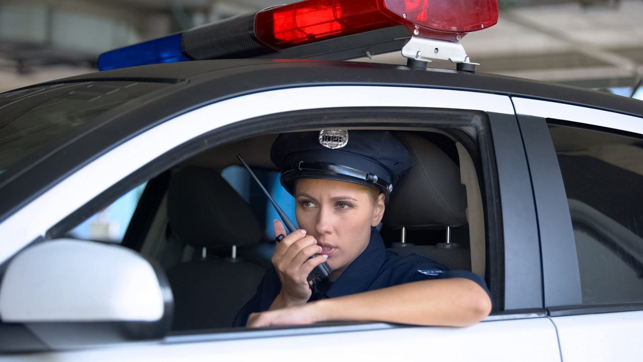 Foto de uma policial segurando um rádio comunicador dentro de uma viatura policial.