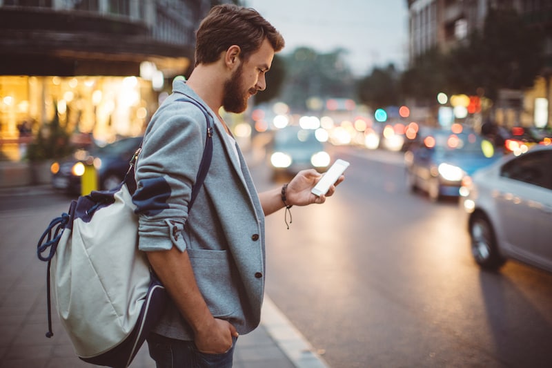 Foto de um homem parado em uma calçada, olhando para a tela de um smartphone. À frente dele, há uma rua com vários carros trafegando.