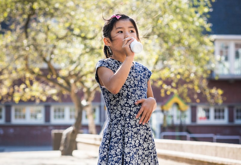 Foto de uma menina, ao ar livre, bebendo um líquido de dentro de uma garrafa. 