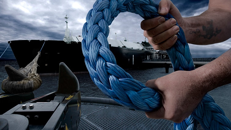 Foto de duas mãos segurando uma corda grossa. Ao fundo, há um navio no mar.