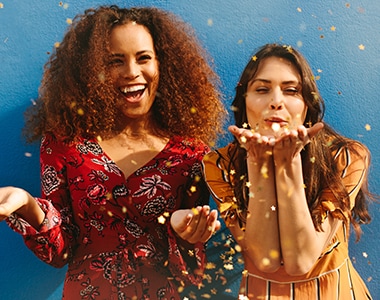 Attractive young women blowing glitters against blue background. Mixed race female friends having fun with glitters.